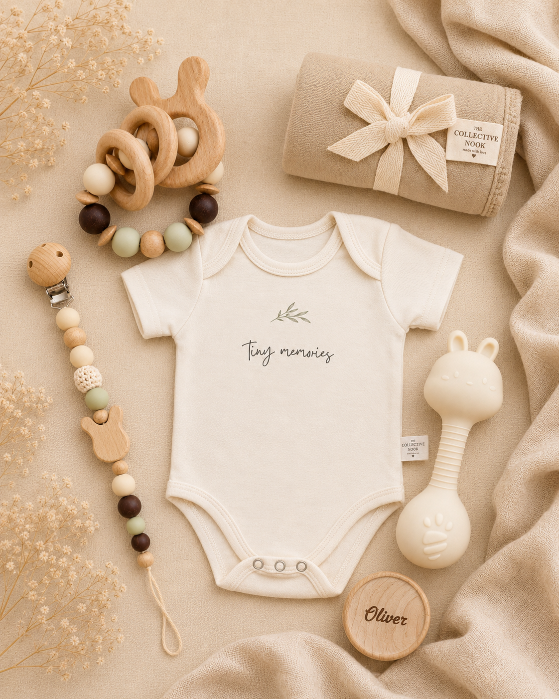 A styled flat-lay of baby accessories on a beige fabric background, featuring a cream short-sleeve bodysuit with 'tiny tumblers' printed on the chest, a wooden teething ring, a wooden bead pacifier clip, a cream bunny teether, and a beige blanket wrapped in a ribbon.