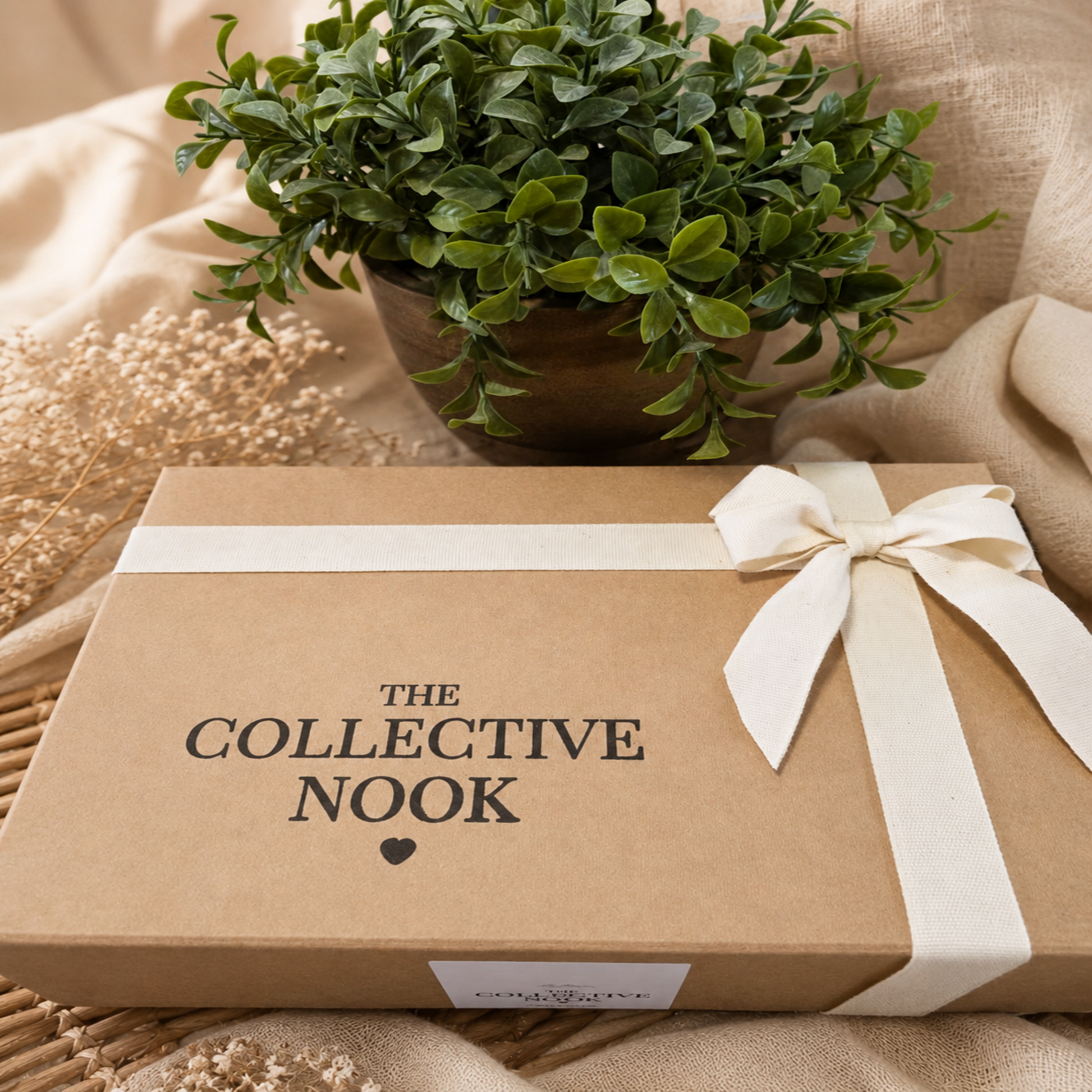 A brown kraft gift box with a cream ribbon and bow, printed with the text 'THE COLLECTIVE NOOK' and a small heart, styled on a beige fabric surface with a potted green plant in the background.