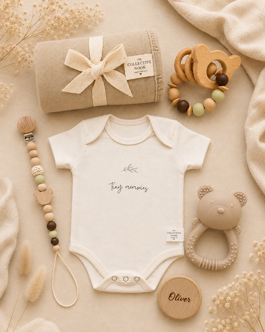 A styled flat-lay of a baby gift set on a beige fabric background, featuring a cream short-sleeve bodysuit with 'tiny names' printed on the chest, a rolled beige blanket with a ribbon and 'The Collective Nook' label, wooden bead teething rings, a wooden bear rattle, and a wooden name plaque.