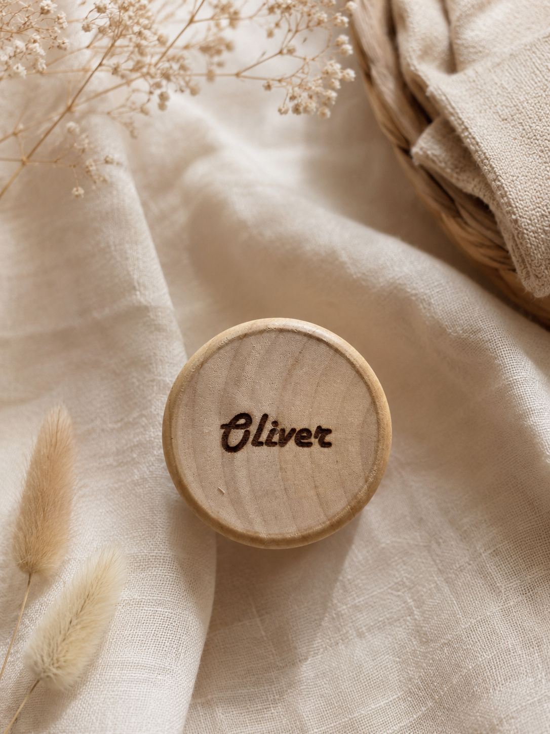 A round wooden keepsake box lid engraved with the name 'Oliver' in dark script, styled on draped beige linen fabric with dried flowers and a woven basket.